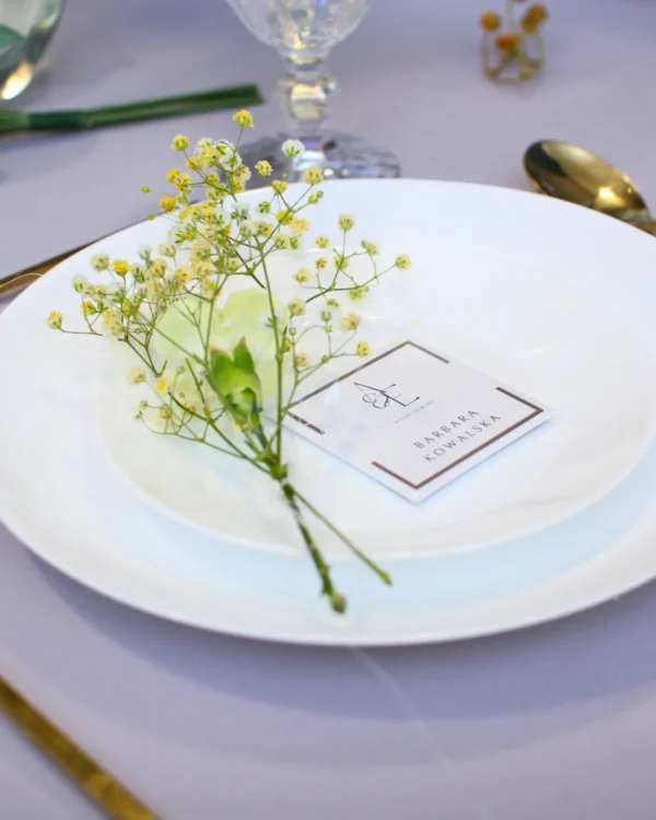 Magnetyczna winietka weselna z imieniem gościa na białym talerzu, delikatne kwiaty i elegancka złota zastawa