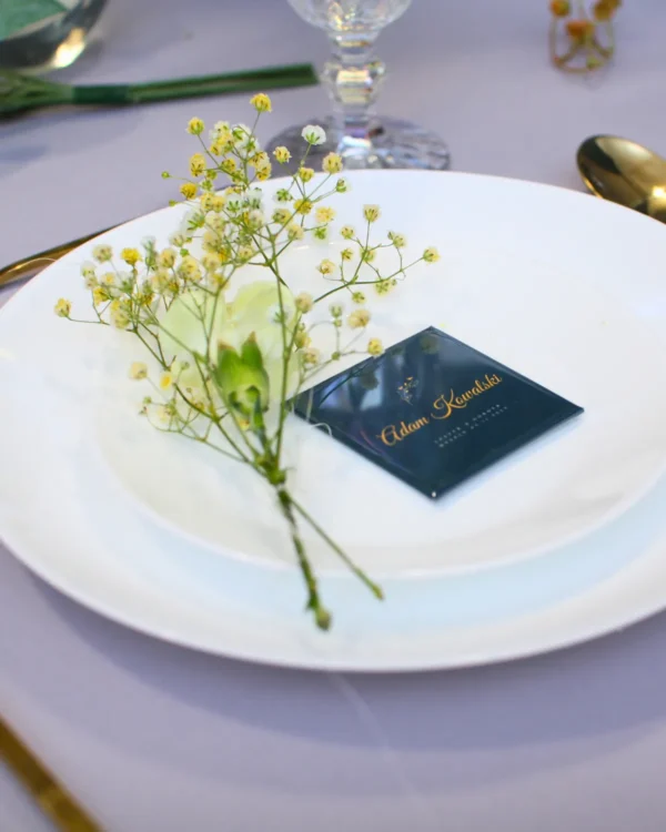 Magnetyczna winietka weselna z imieniem gościa na granatowym tle, białe talerze, kwiaty i elegancka złota zastawa