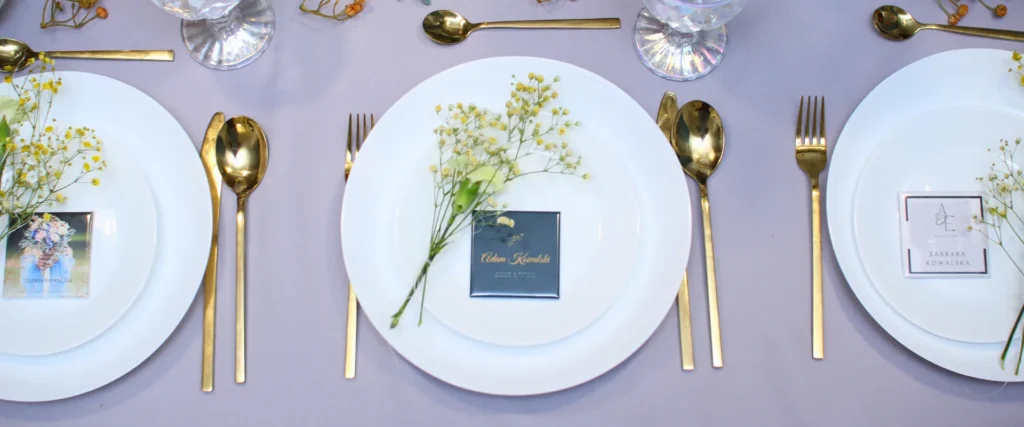 Elegancka aranżacja stołu weselnego z białymi talerzami, złotymi sztućcami, delikatnymi kwiatami i różnymi wariantami winietek magnetycznych dla gości.