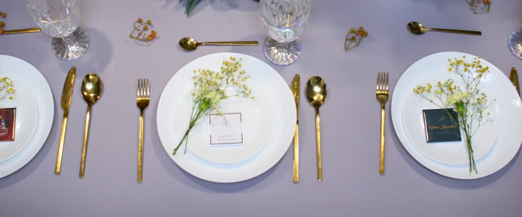 Elegancka aranżacja stołu weselnego z białymi talerzami, złotymi sztućcami, kryształowymi kieliszkami, delikatnymi kwiatami i winietkami magnetycznymi dla gości.