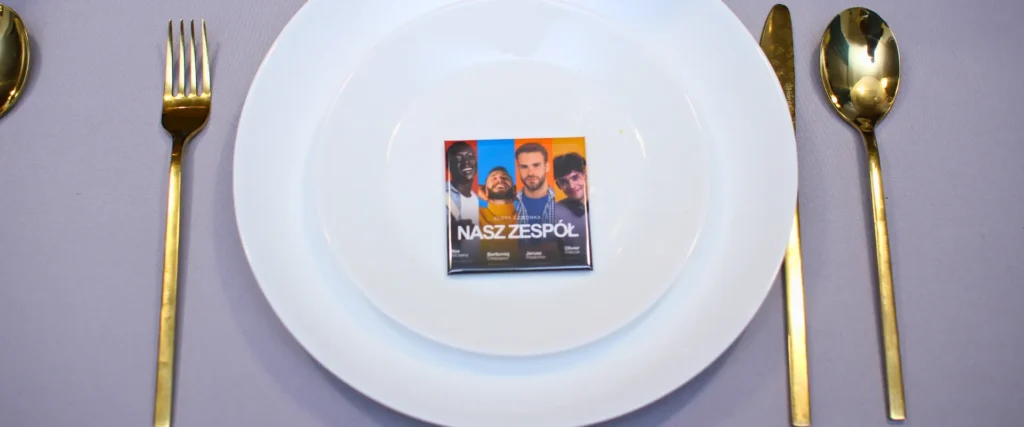 Winietka magnetyczna z grafiką „Nasz zespół” ułożona na białym talerzu, otoczona złotymi sztućcami na elegancko nakrytym stole.