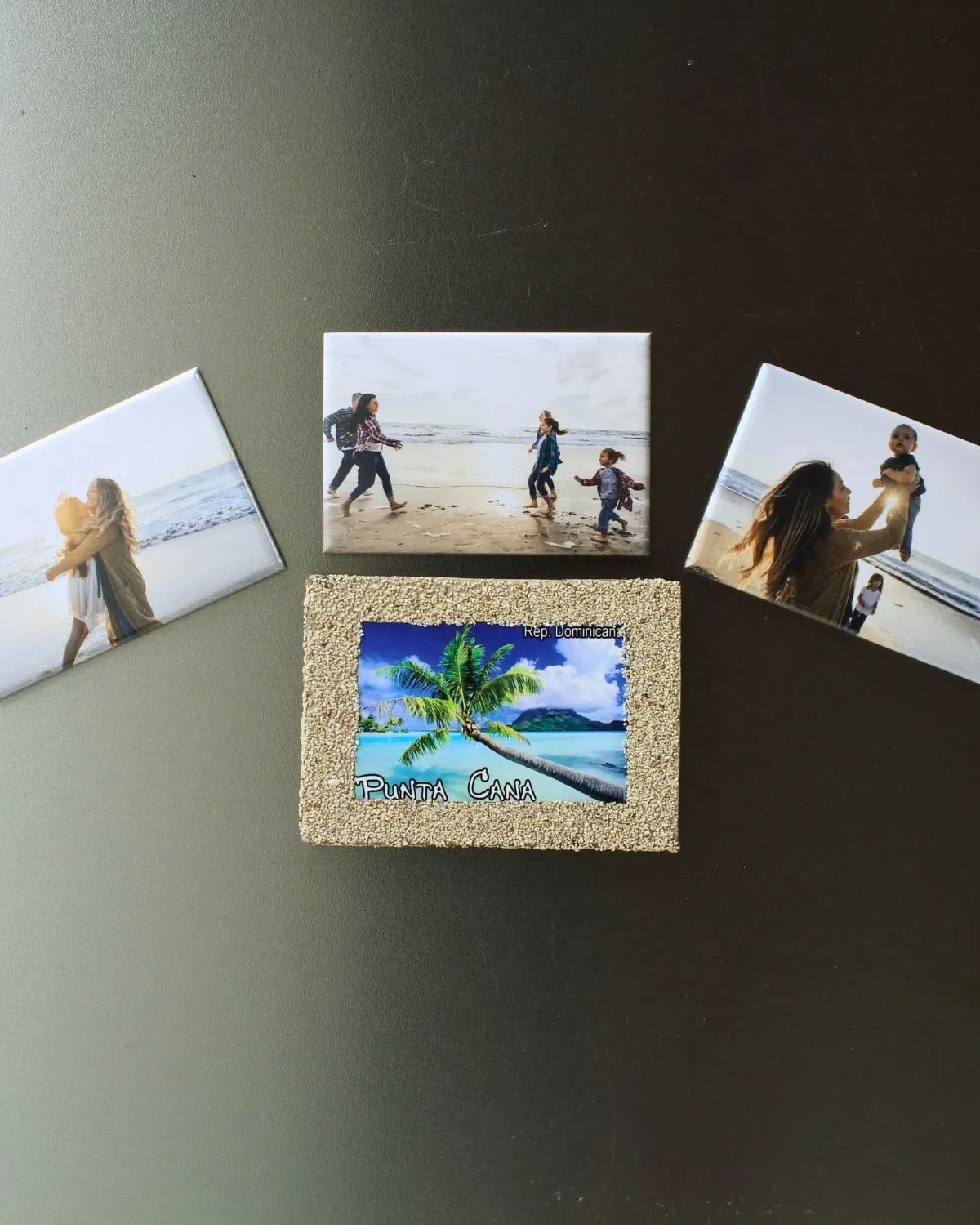 Trzy prostokątne foto magnesy z rodzinnymi zdjęciami z plaży zostały przypięte do metalowej powierzchni wokół pamiątkowego magnesu „Punta Cana”. Kompozycja pokazuje, jak personalizowane foto magnesy można łączyć z magnesami z podróży, tworząc estetyczną i pełną wspomnień dekorację lodówki lub tablicy magnetycznej.